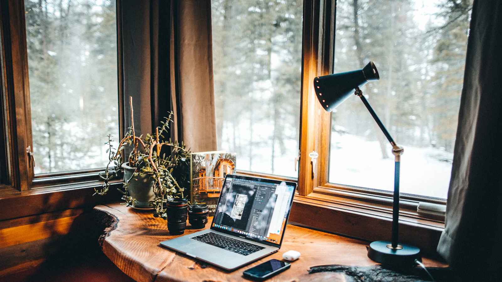 A wooden desk in a snowy window nook with a laptop, a black desk lamp and a potted plant.